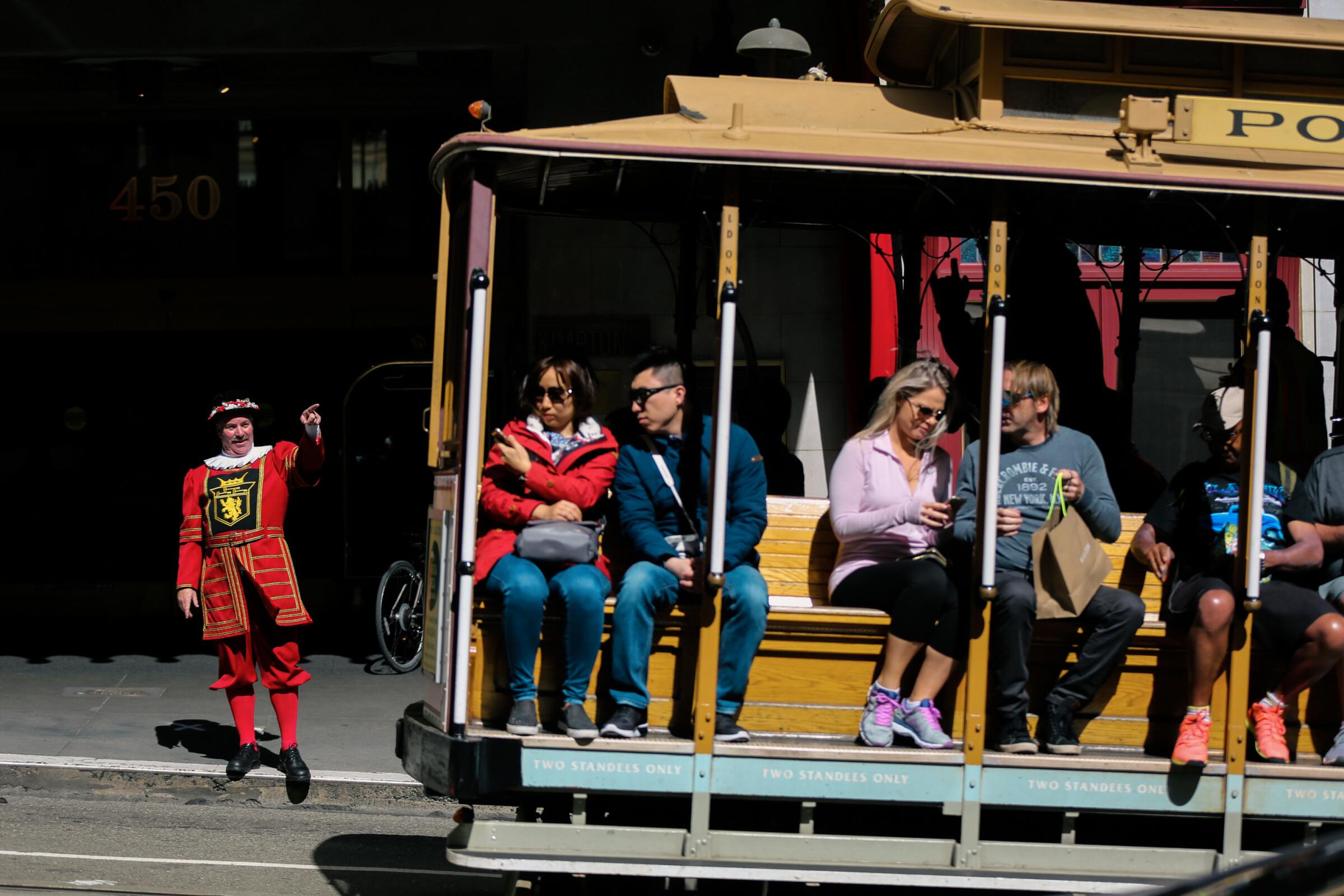 A Sir Francis Drake hotel doorman dressed in traditional beefeater garb points at a passing cable car.