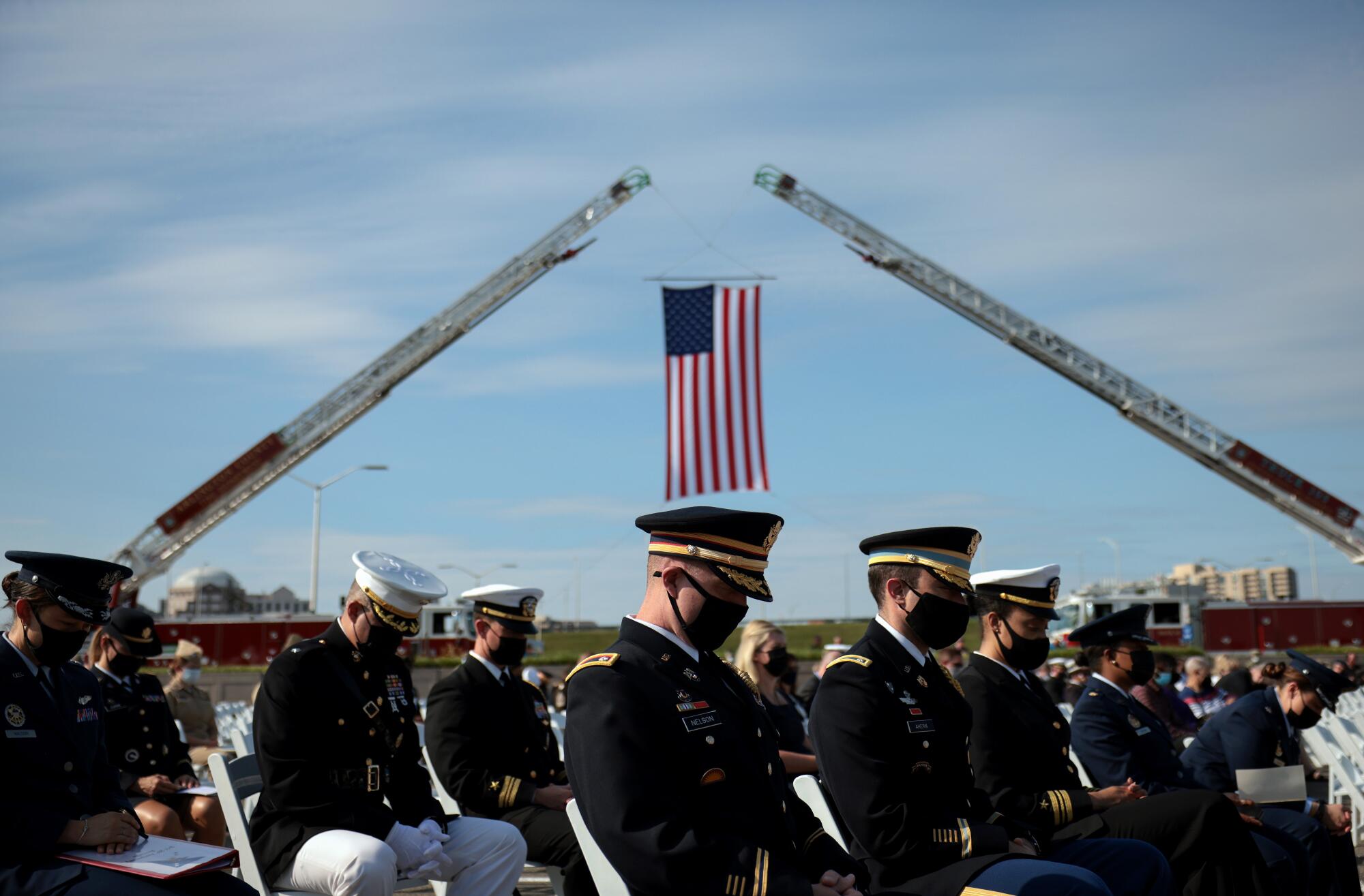 U.S. service members sit in chairs while a U.S. flag hangs from two cranes behind them