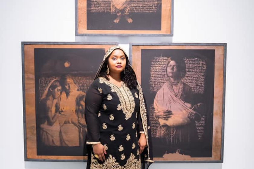 A woman in a intricate embroidered outfit and a head covering stands in front of three images of Indian women
