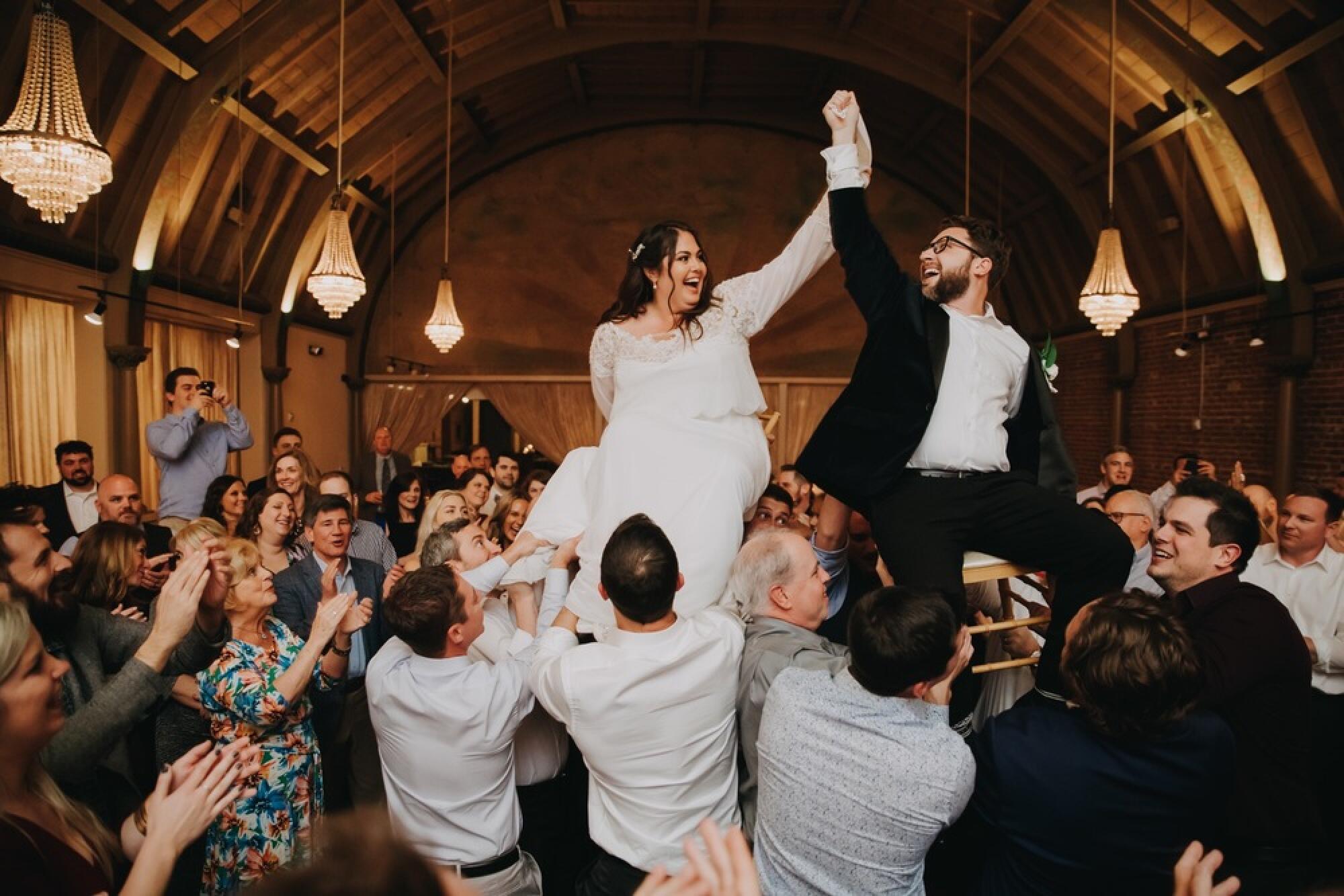 A crowd lifts a bride and groom seated on chairs at their wedding.