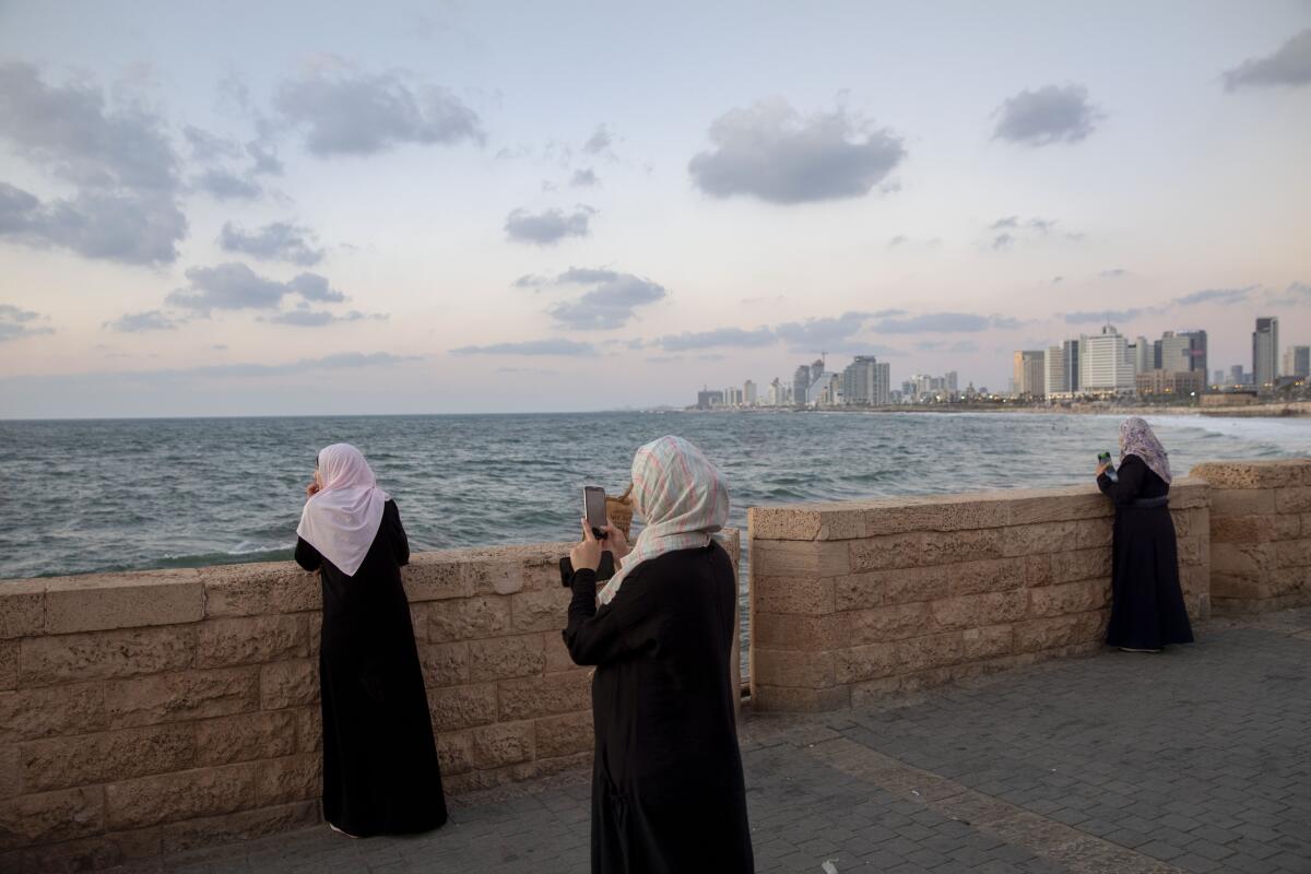 Women in headscarves and robes stand on a waterfront walk facing the sea.