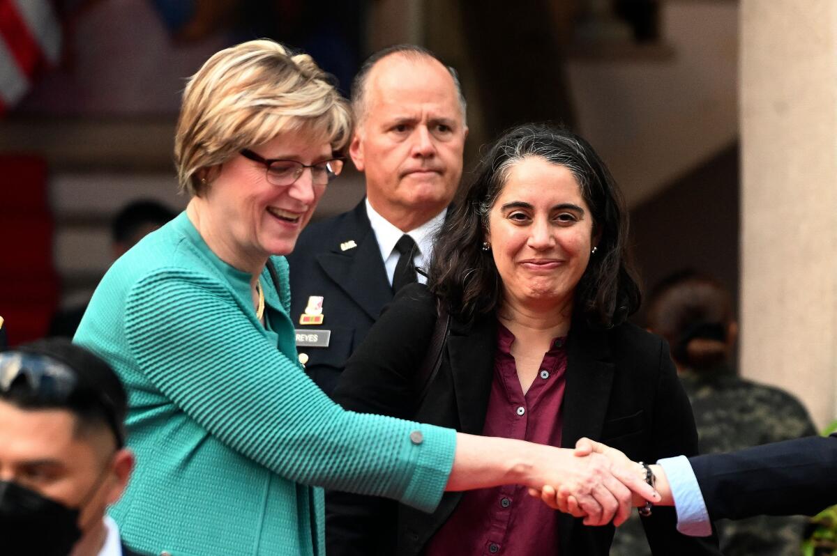 A woman in glasses and a turquoise jacket shakes hands with an unseen person as other people look on.