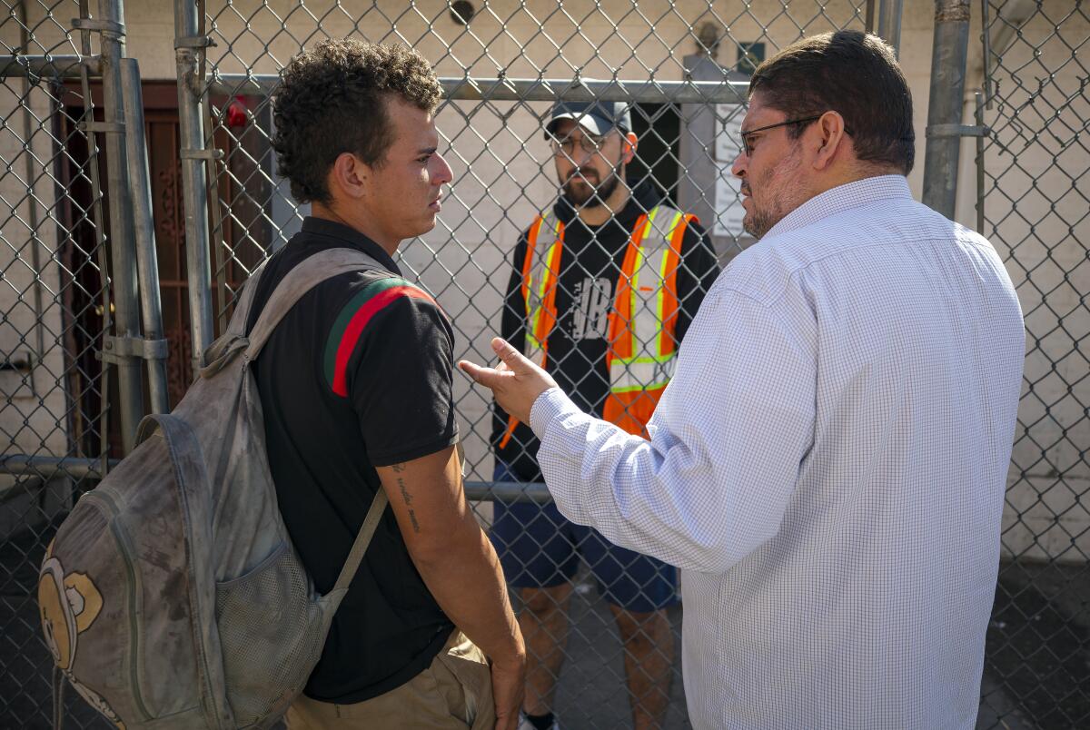 A man carrying a travel bag talks with another man in front of a chain link fence while a man in a safety vest looks on. 