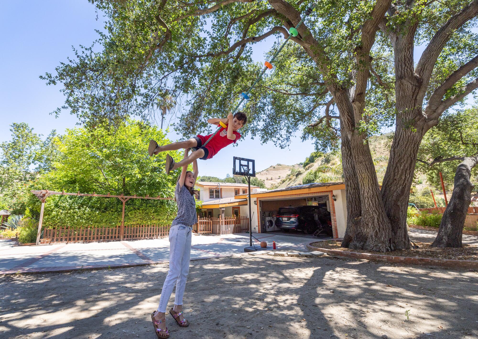 Two children, one suspended on a rope swing, play in a large yard.