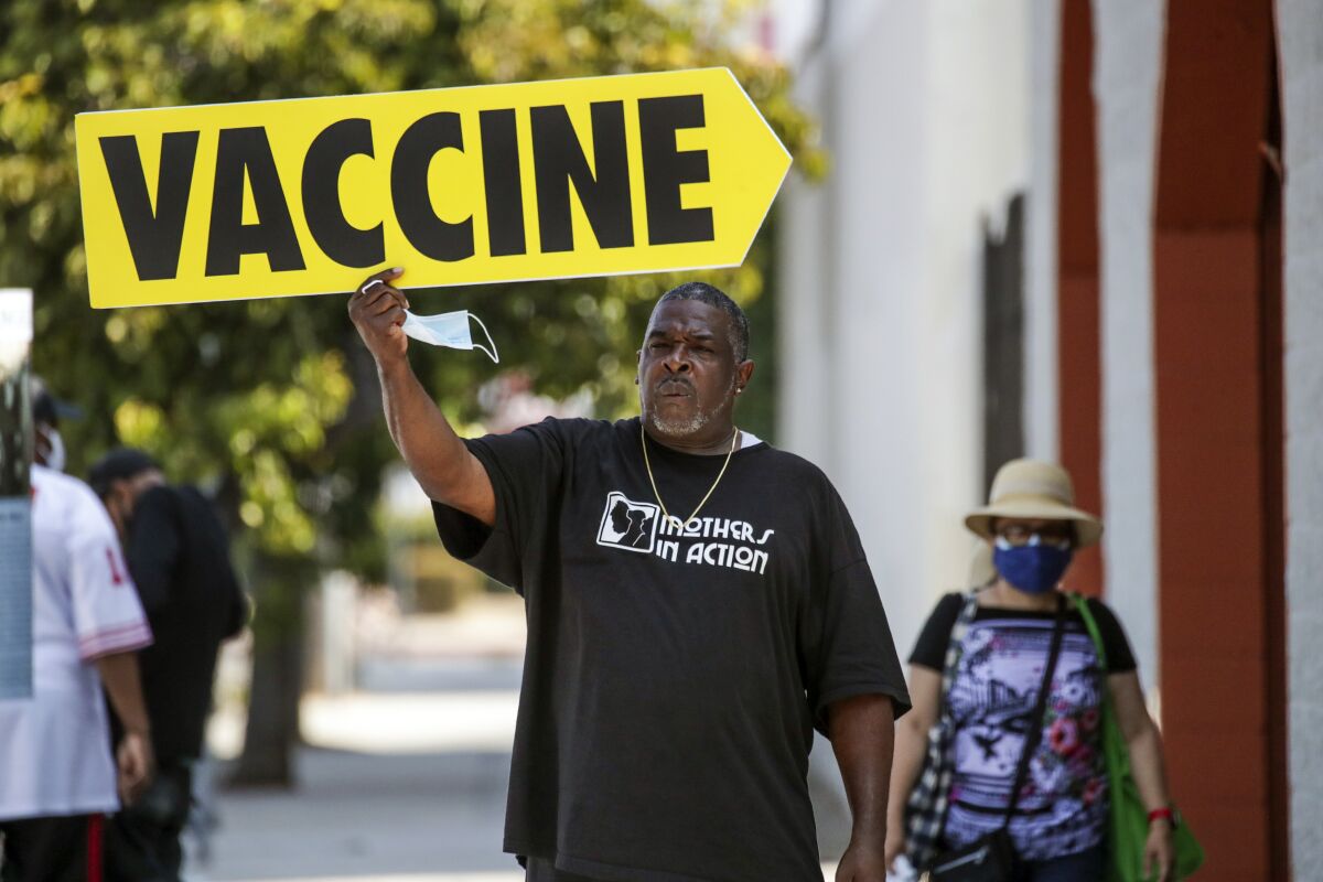 A man in a "Mothers in Action" T-shirt holds an arrow-shaped sign reading "Vaccine"