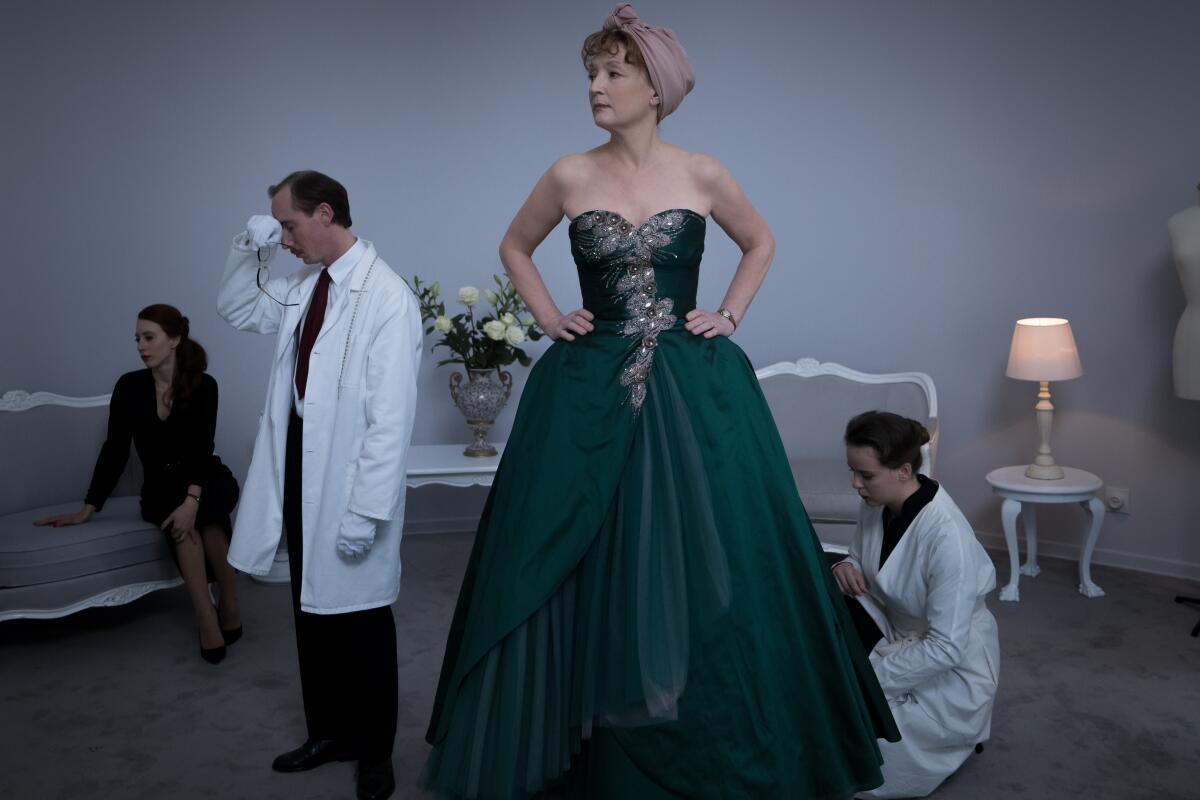 A woman stands in a strapless gown. A woman kneels nearby as a man in lab coat and gloves presses his hand to his brow.