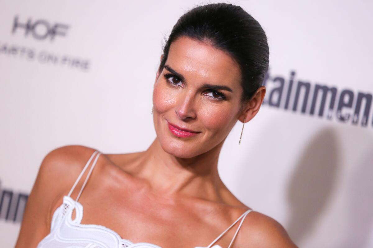 A slightly tilted view of Angie Harmon posing in a white, thin strapped gown with her hair pulled back