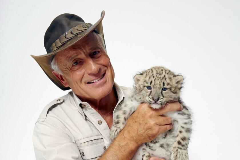 Jack Hanna wears a wide-brimmed hat and hold a snow leopard cub while sitting on the floor