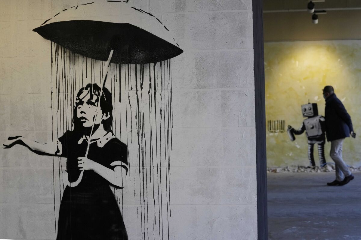 "The Umbrella girl" a reproduction of a mural by British artist Banksy is seen as a visitor walks past in background during the unveiling of the "The World of Banksy, The Immersive Experience" exhibition, in Milan, Italy, Thursday, Dec. 2, 2021. An exhibition of 130 works by British street artist Banksy opens Friday in a gallery space inside Milan's Central train Station. The exhibition unveiled on Thursday includes 30 never before seen works by Bansky and highlights pieces by young unknown artists from all over Europe. (AP Photo/Luca Bruno)