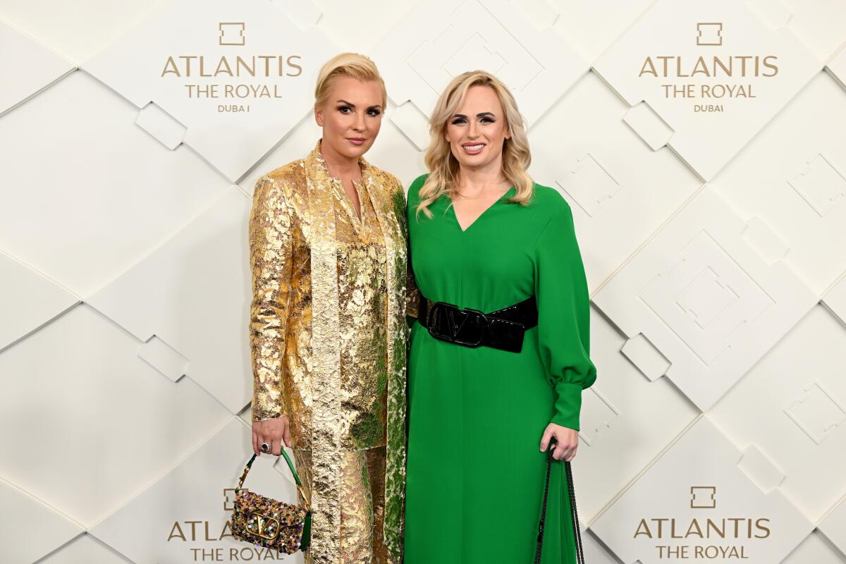 Two blond women, one in a shimmering gold suit and another in a green dress and big black belt, stand in front of a backdrop