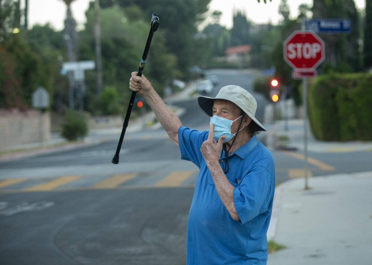 Charles Dirks raises his cane and points to his face from the side of the road to remind a motorist to wear a mask