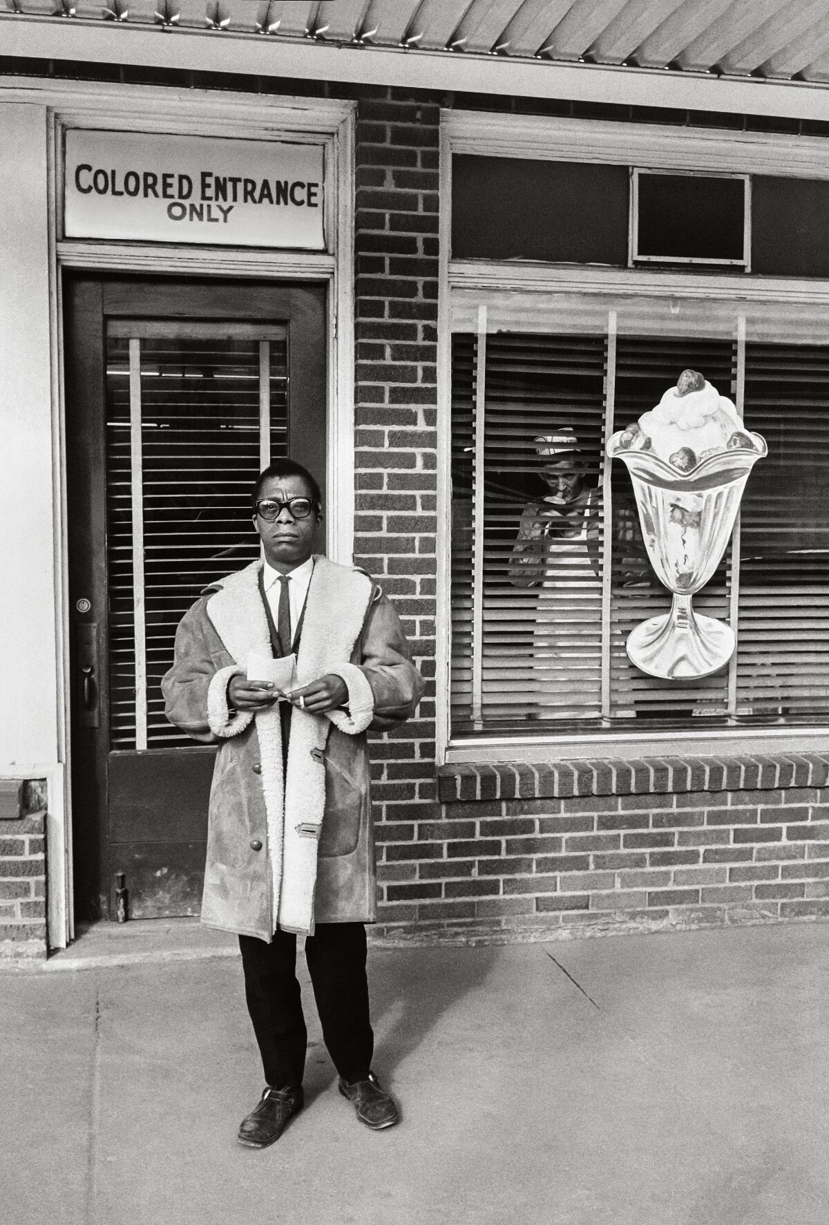 A man in a coat stands in front of an ice cream shop and a sign that reads, "Colored entrance only."