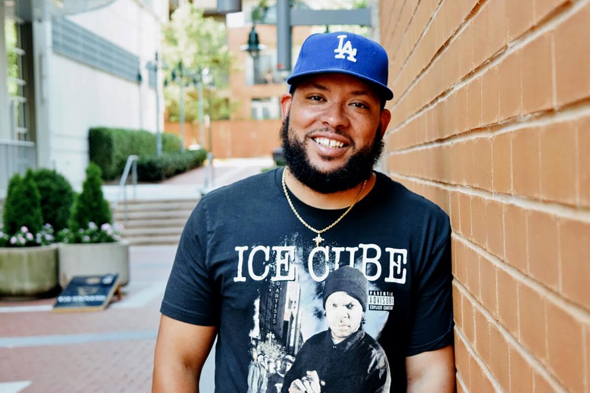 A man wearing a Dodgers cap and an Ice Cube T-shirt leans against a brick wall.