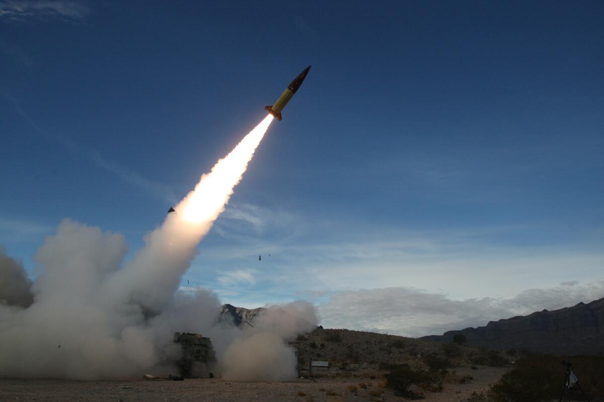 A missile soars skyward in a desert setting, leaving a fiery contrail and clouds of smoke on the ground