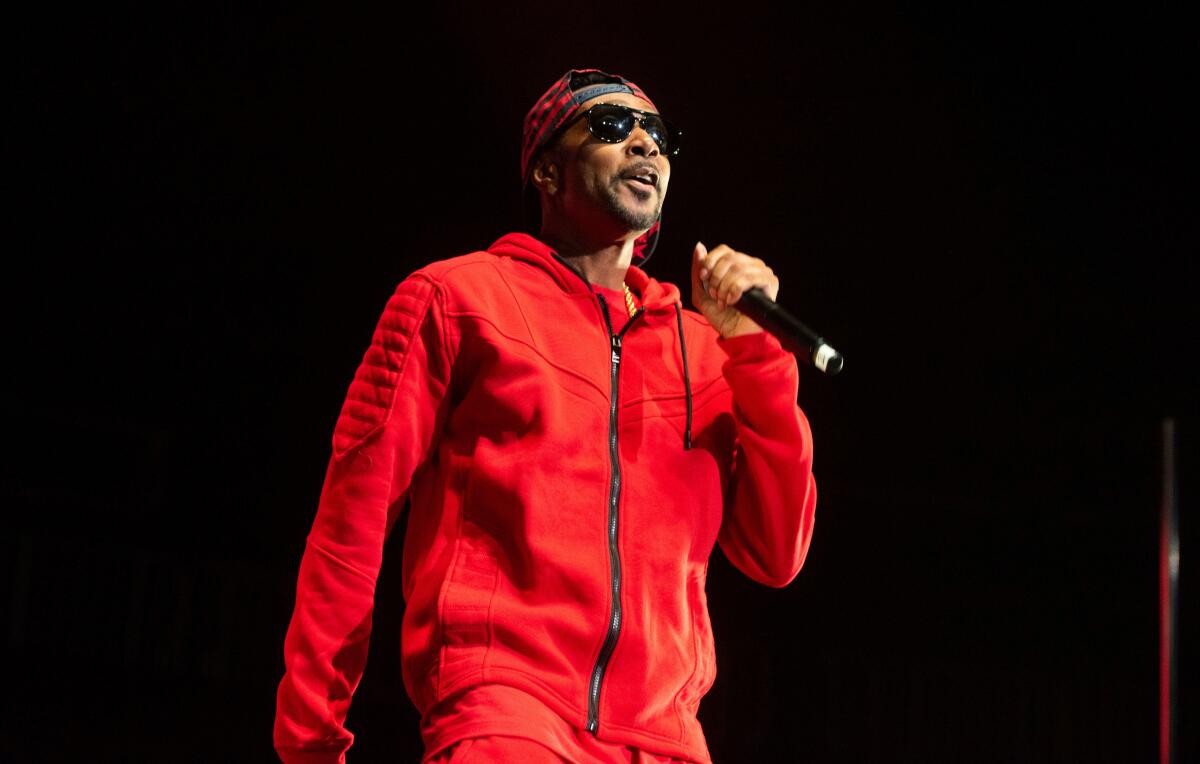 Krayzie Bone raps into a mic on a dark stage in a red tracksuit, dark glasses and backward cap