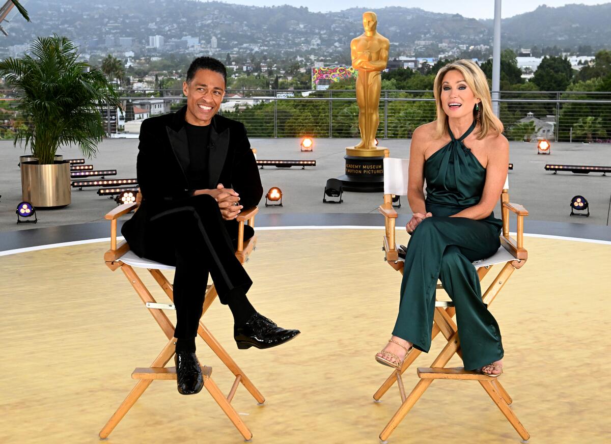 T.J. Holmes wears all black and Amy Robach wears a green halter jumpsuit as both sit in director's chairs on location