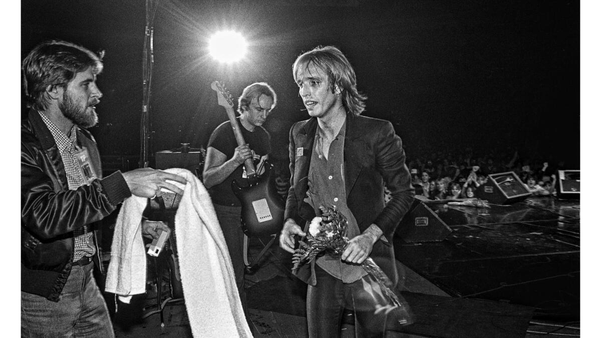 Black and white photo of Tom Petty walking off a concert stage with flowers in his hand as a man hands him a towel