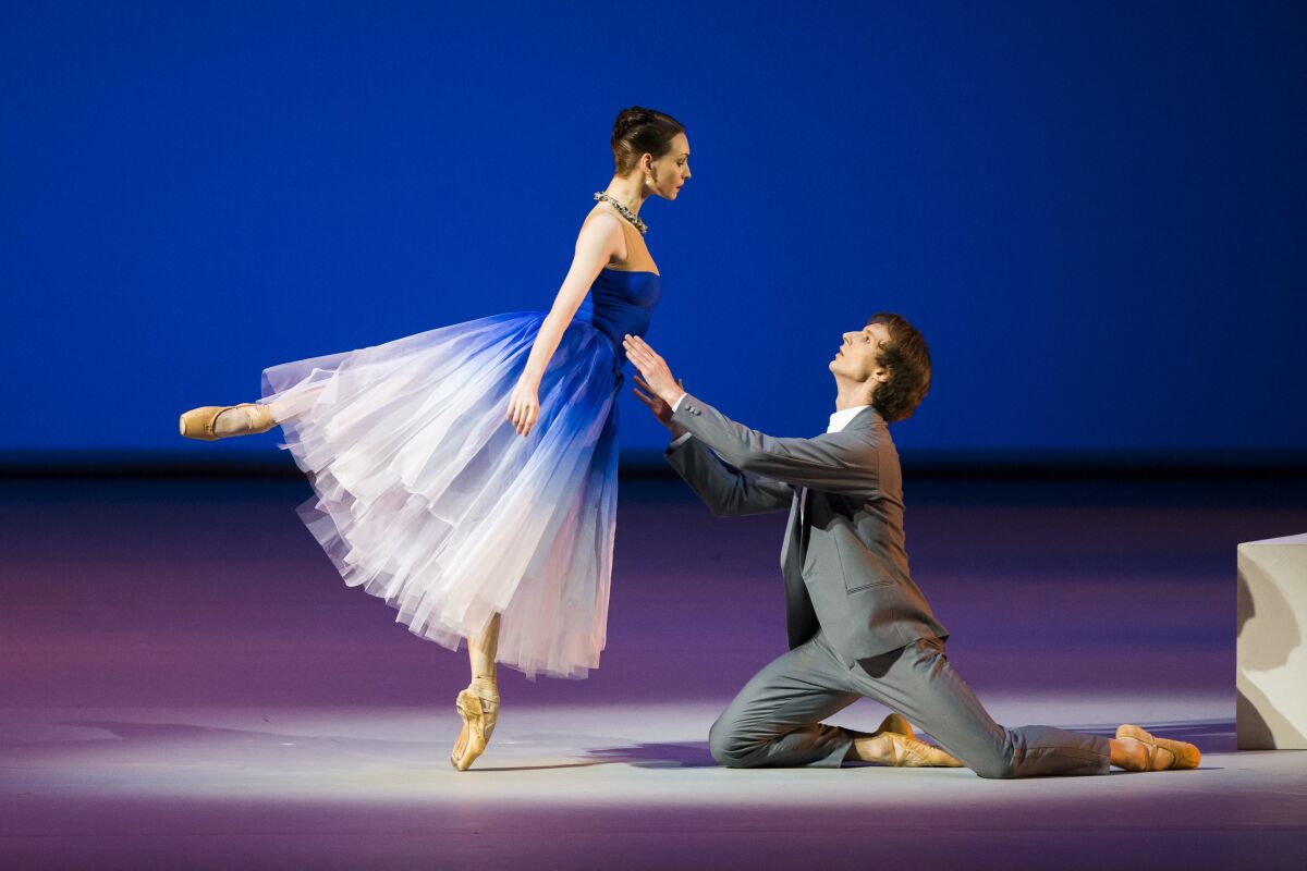A ballerina in a blue and white gown leans into a male dancer, who kneels on the stage.