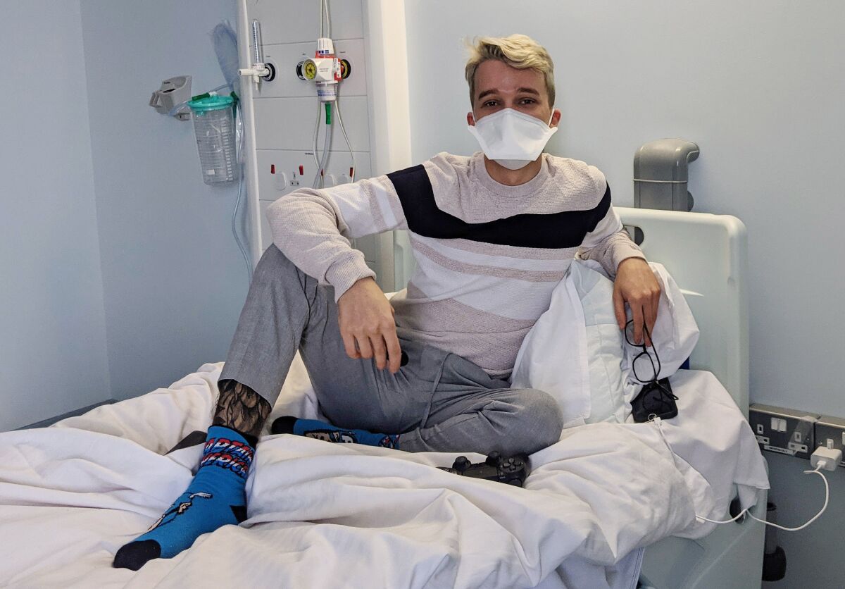 A young man in a face mask sits fully clothed in a hospital bed. His posture is casual and relaxed. 
