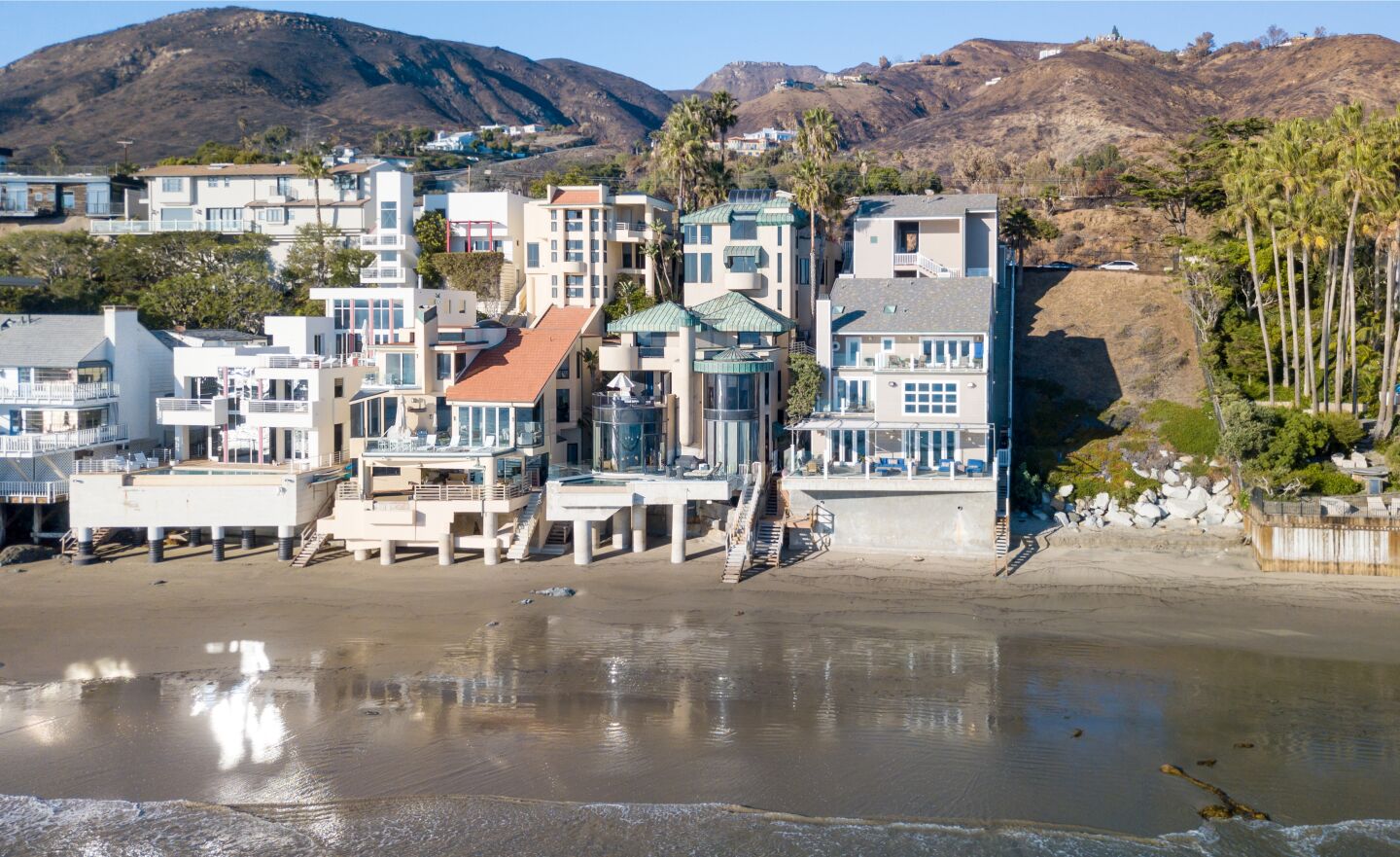 The beachfront home is steps from the ocean along the beach with other homes and hills behind them.