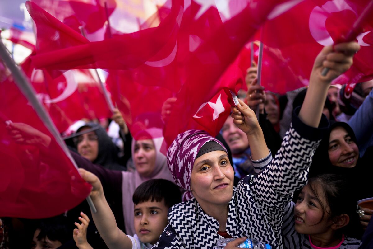 A woman stands close the the camera, smiling, as she and a crowd of people wave Turkish flags
