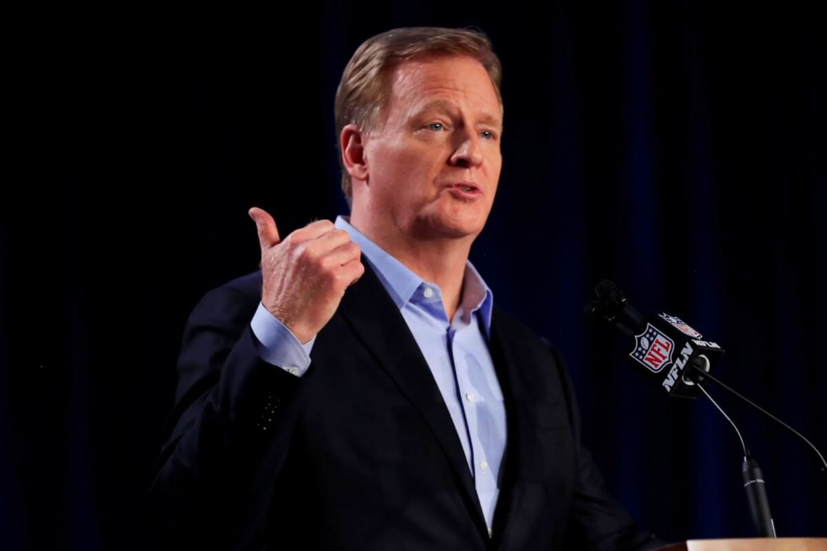 NFL Commissioner Roger Goodell holds his hand in the air with his thumb out while he's at a microphone.