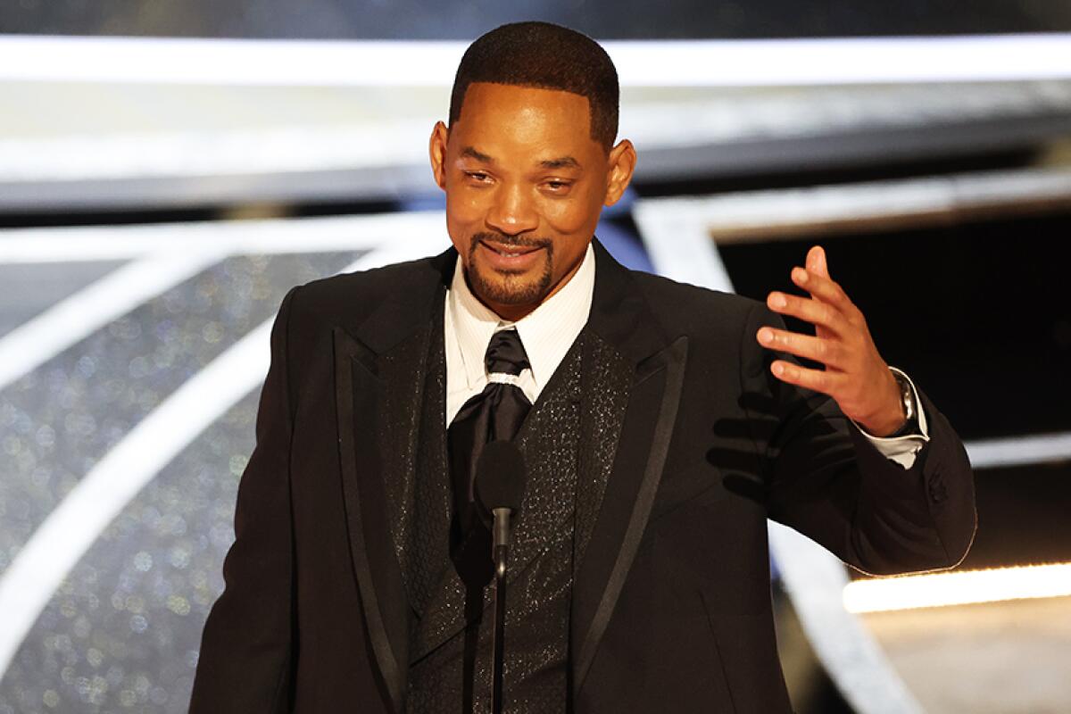 A man in black formalwear smiles and gestures in front of a microphone onstage