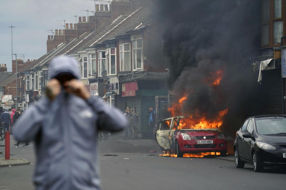 A person with a hoodied pulled tight around his face walks down an English street as a car burns in the background. 