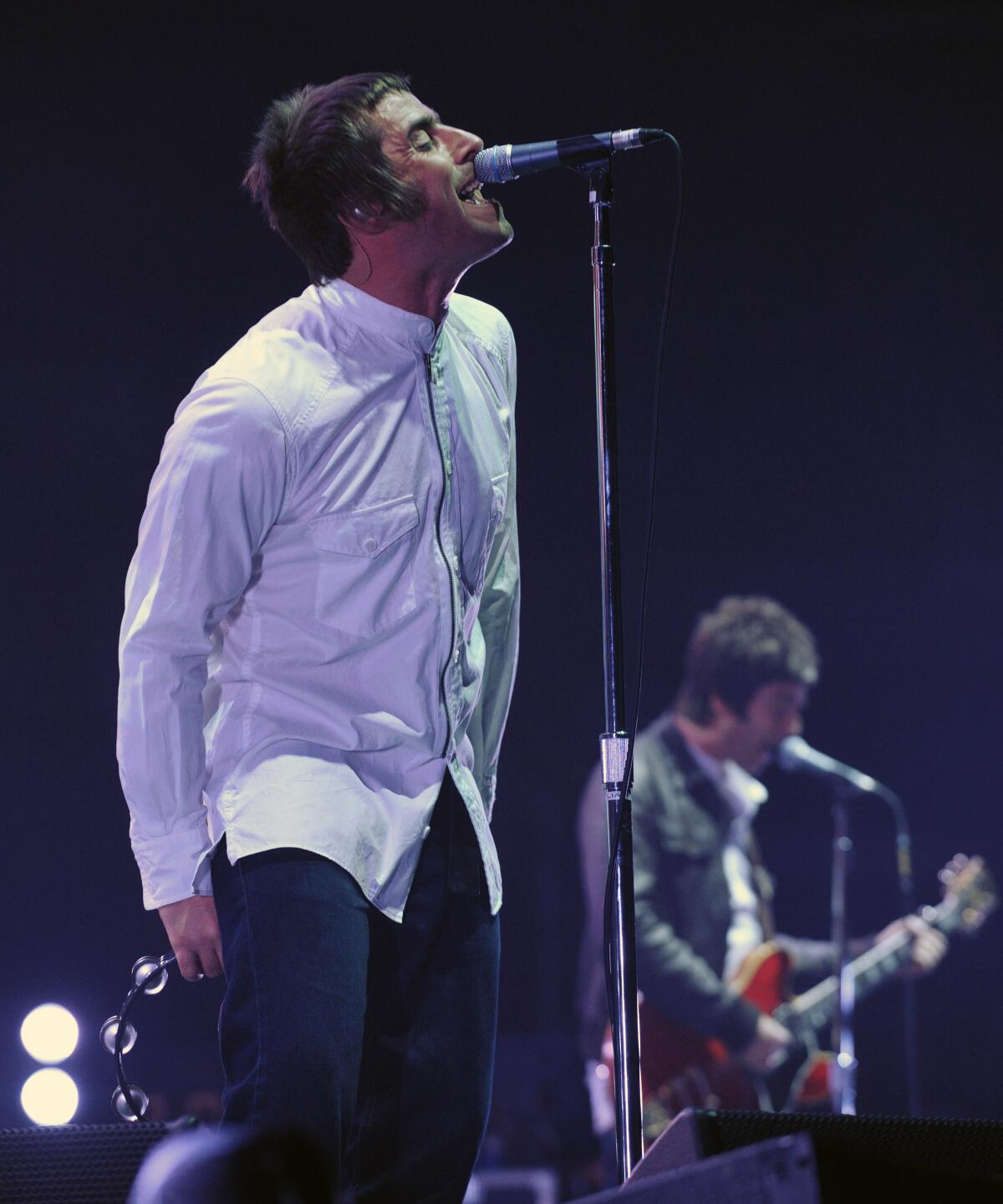 Liam Gallagher singing into a microphone while his brother Noel Gallagher plays guitar onstage