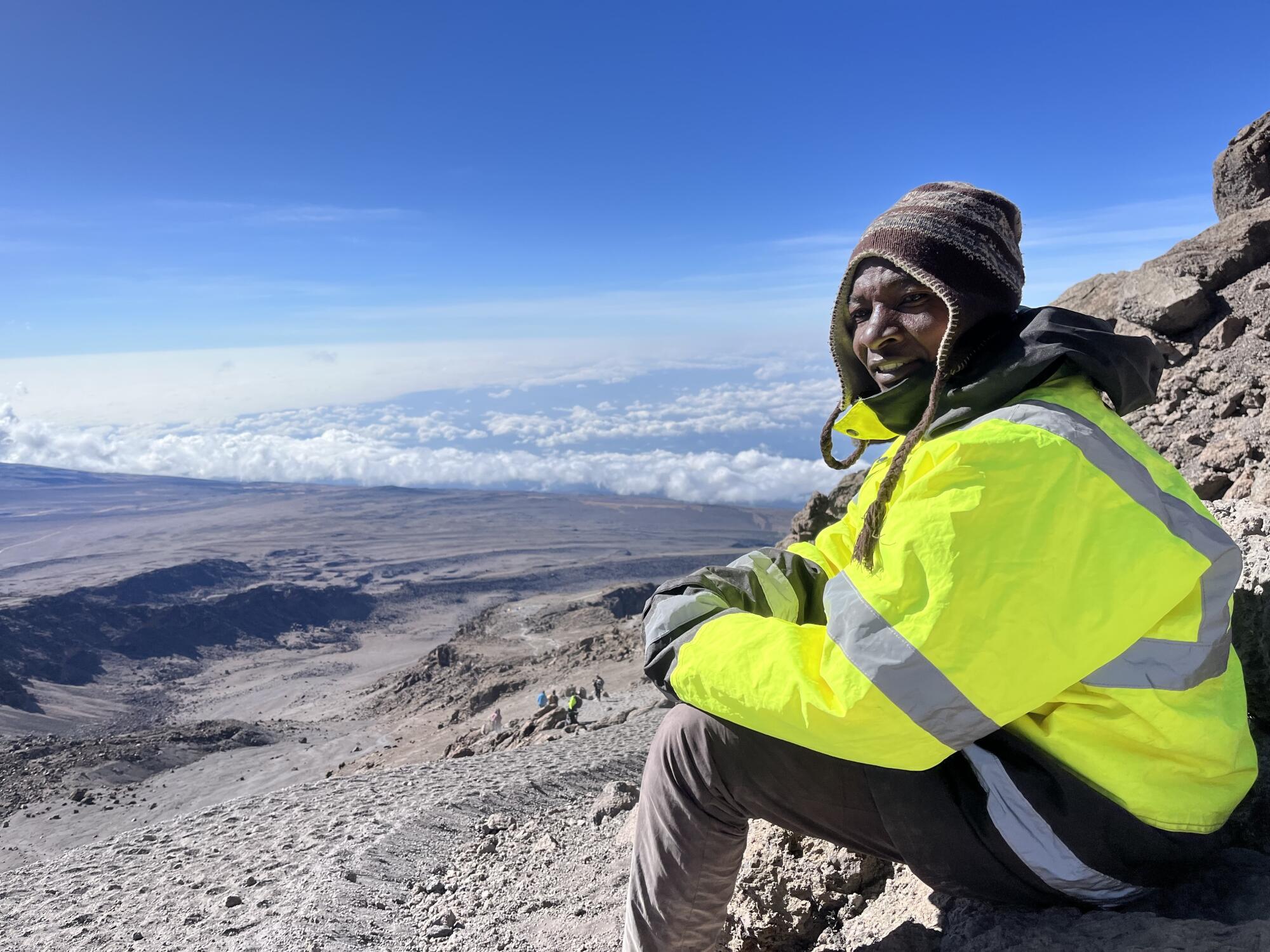 A person in a fluorescent green jacket and knit hat sits amid a brown mountainous landscape 