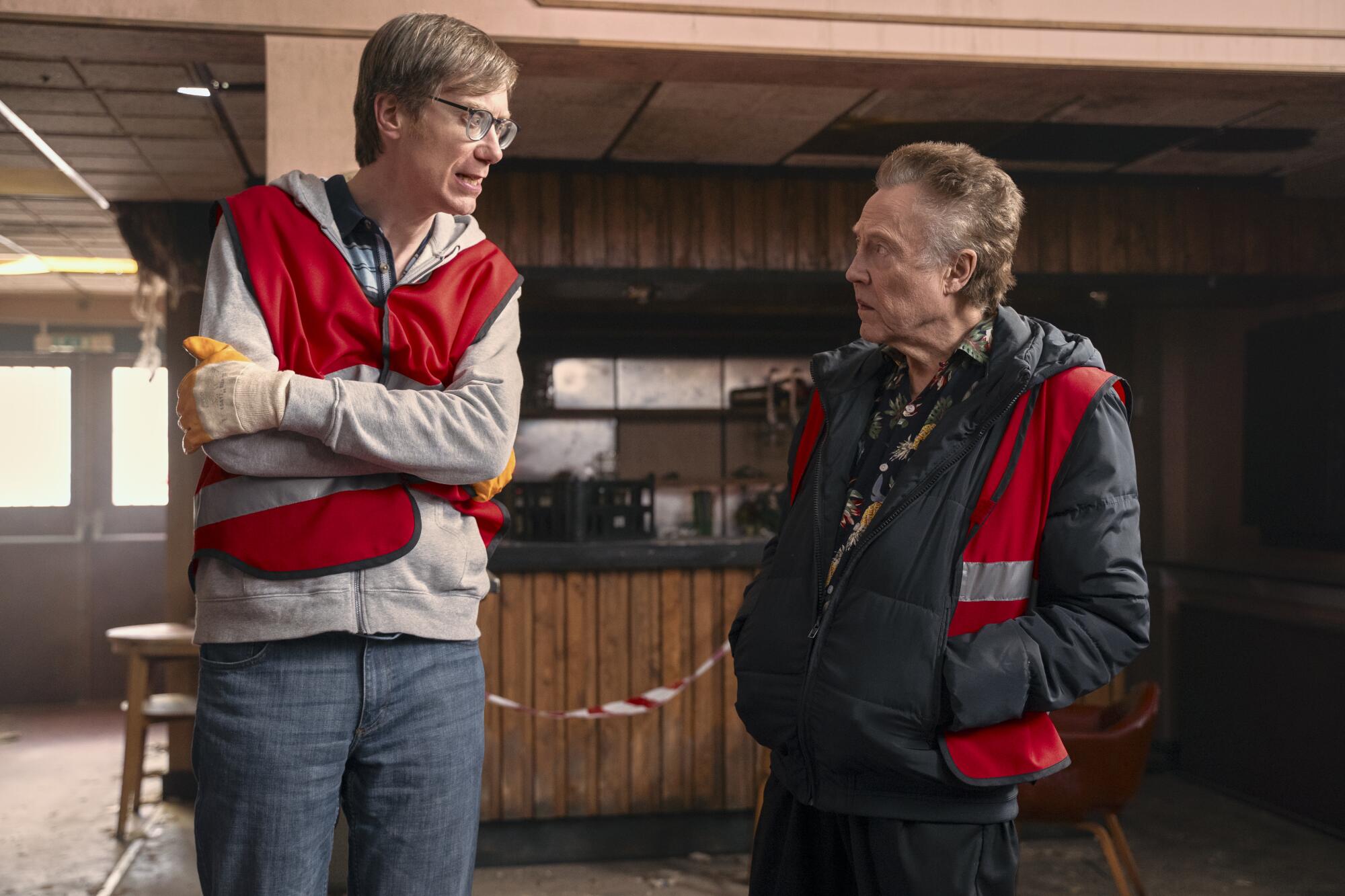 Dos hombres con chalecos reflectantes rojos hablan seriamente en "El proscrito."