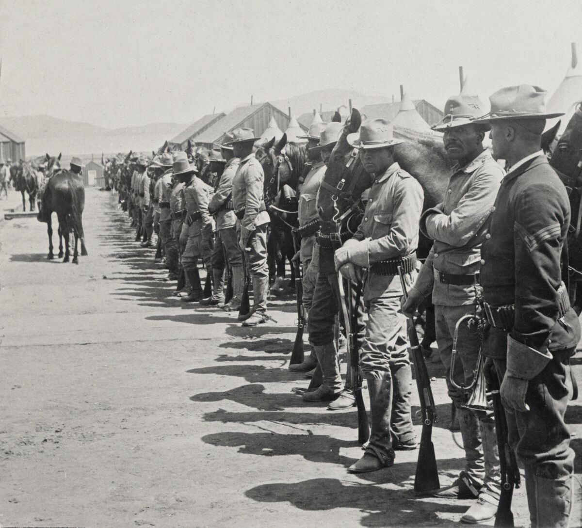 Men in uniforms that include hats and knee-high boots stand in a line holding rifles. Nearby, a soldier leads a horse.