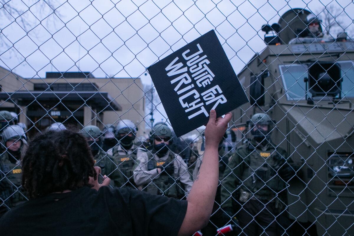 A man holds a sign that says Justice for Wright in front of a chain-link fence with heavily armored police on the other side