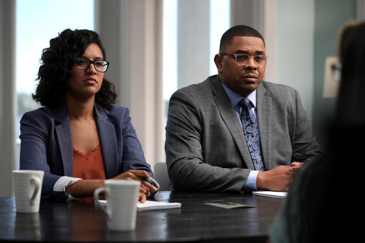 A man and a woman wearing blazers and glasses look at an interview subject.