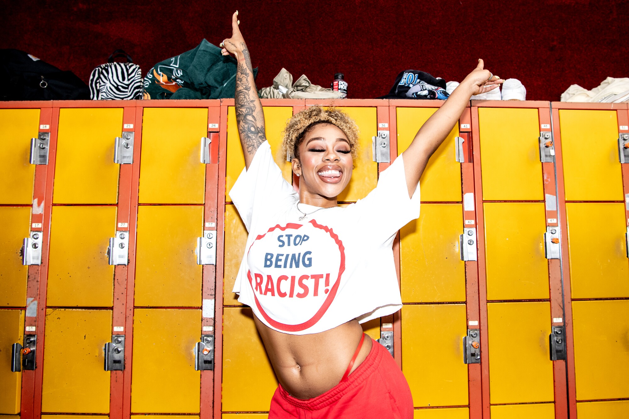 A woman posing raises her arms in jubilation wearing a shirt that says "Stop being racist!"