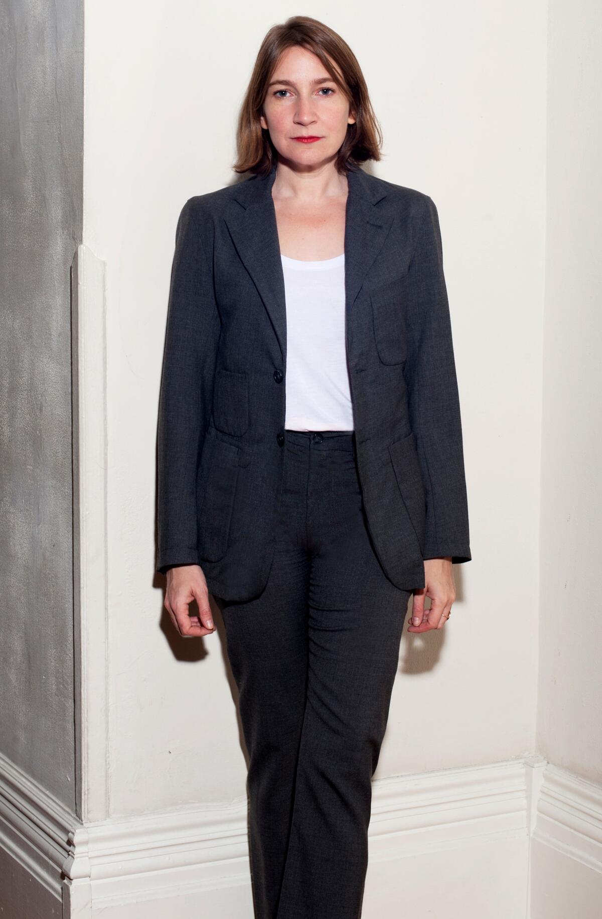 Author Sheila Heti, dressed in a suit, stands in a corner against a white wall with baseboards.