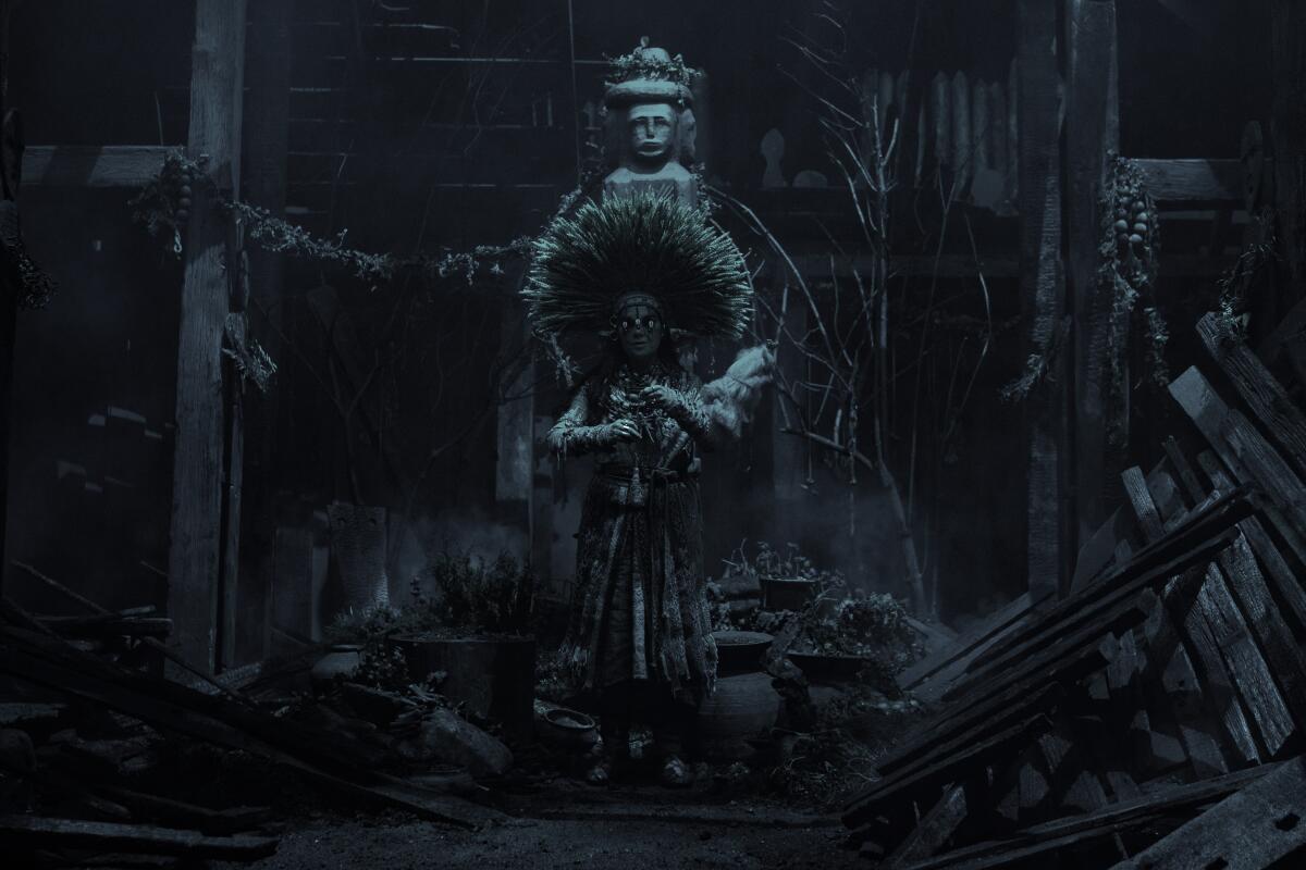 A woman wearing an elaborate headdress stands in a crude hut with an idol behind her. 