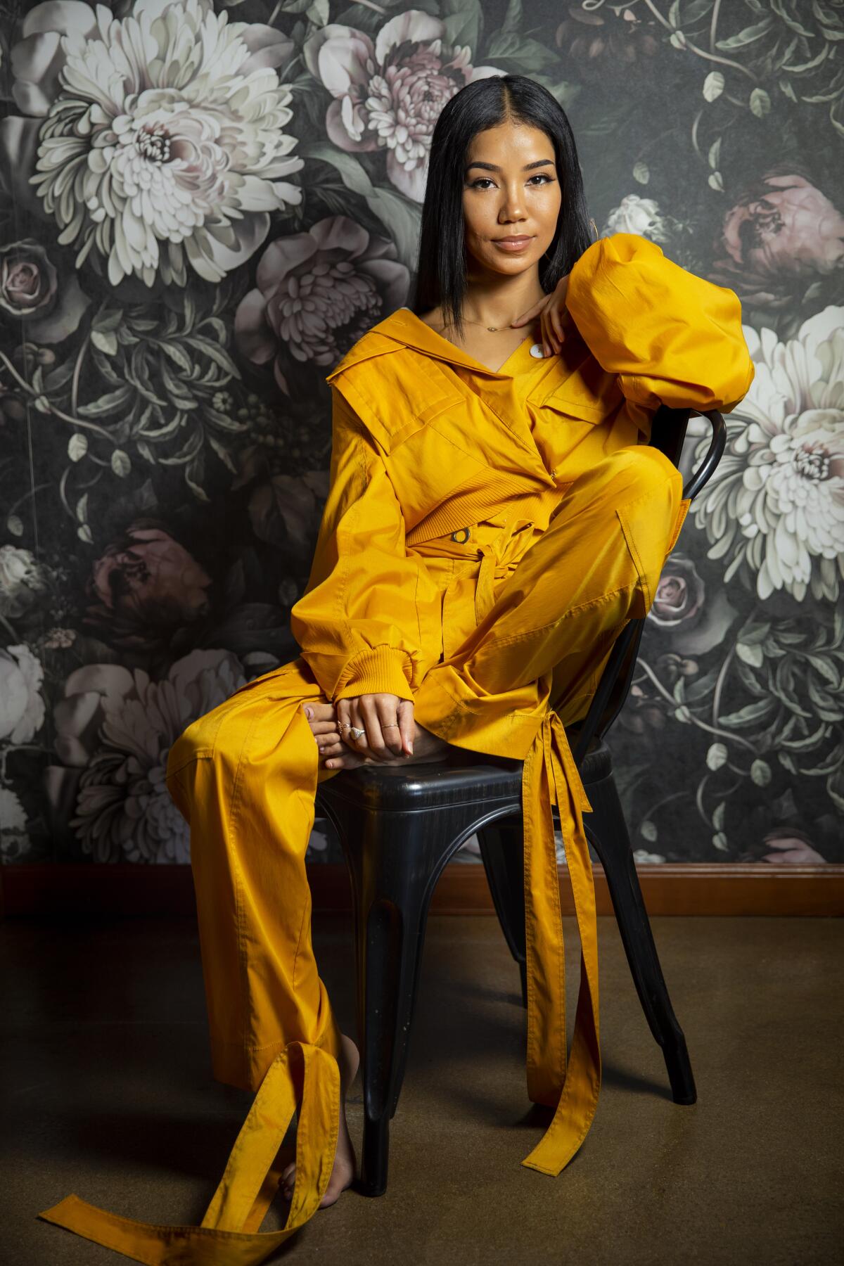 Jhené Aiko, seated and wearing a jumpsuit, poses for a portrait.