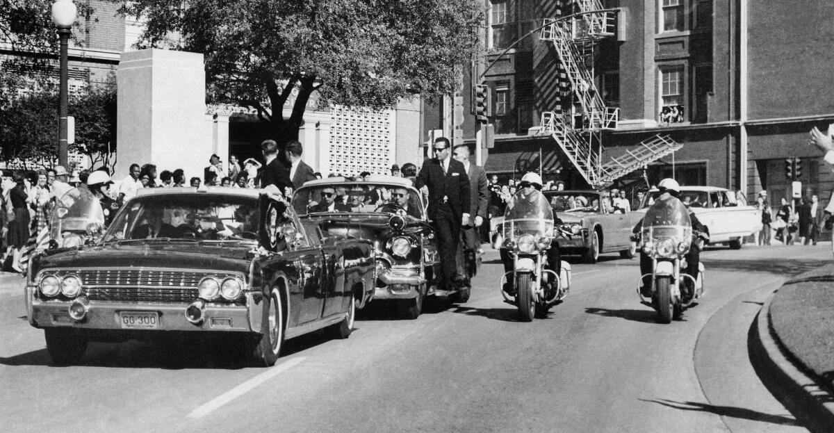 Seen through the foreground convertible's windshield, President John F. Kennedy's hand reaches toward his head within seconds of being fatally shot.