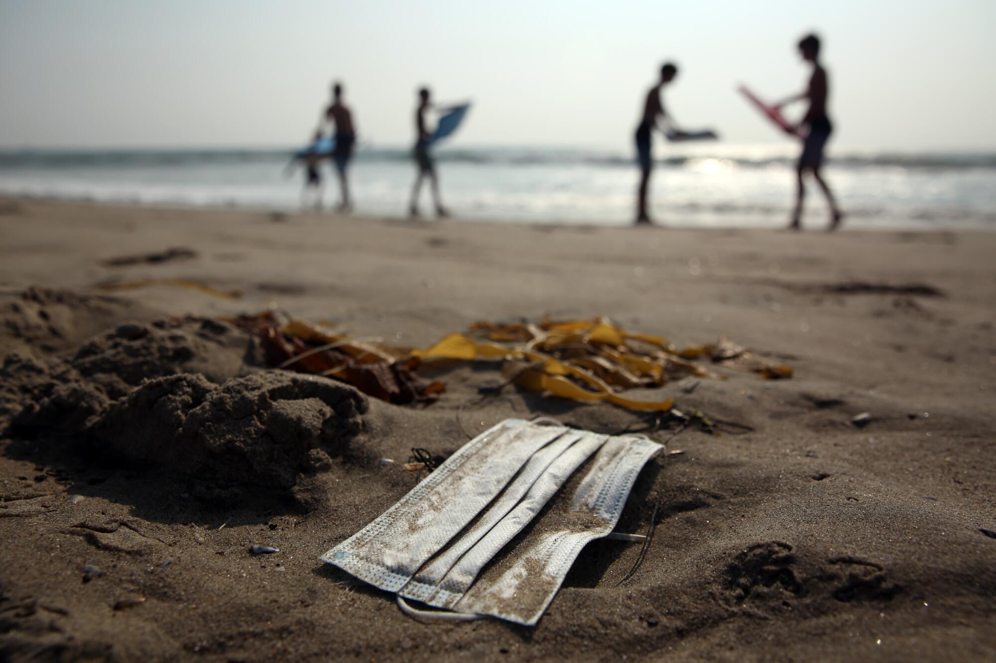 A discarded mask on the beach as people with boogie board stand in the background