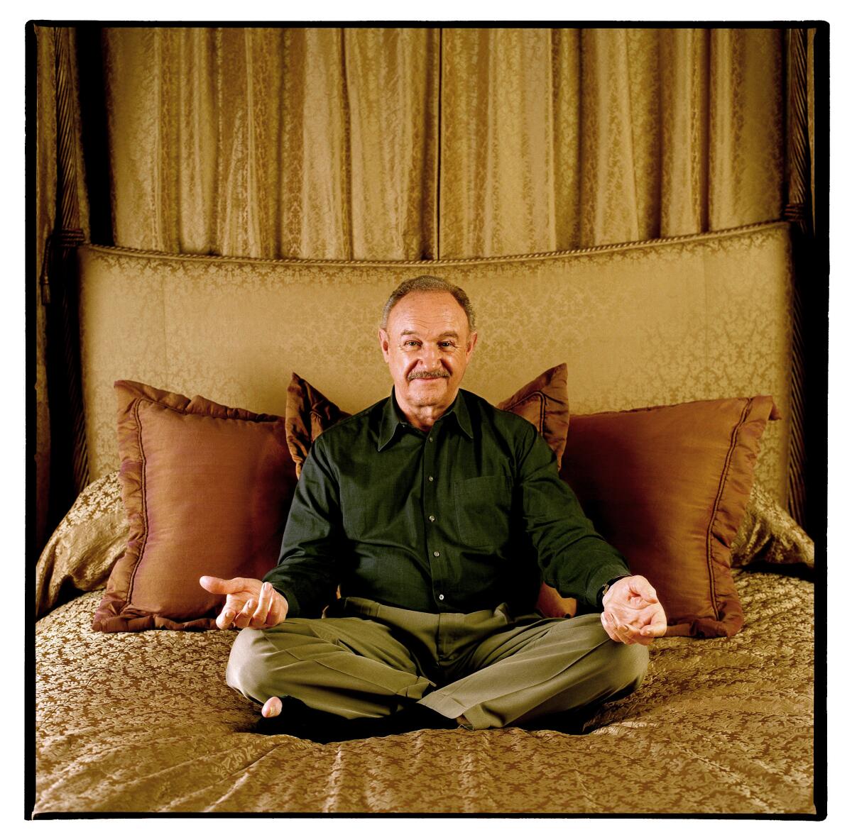 Gene Hackman sits cross-legged connected a furniture with his palms unfastened facing up and resting connected his knees.