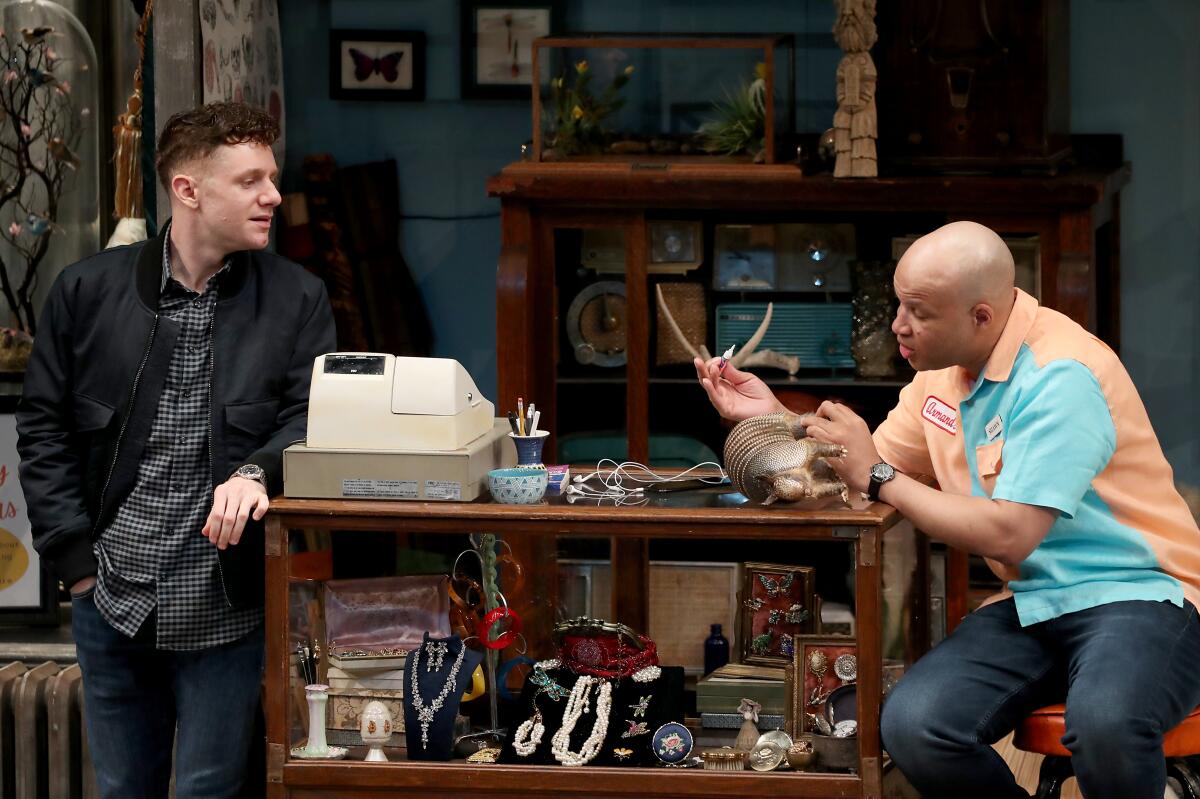 Two men converse on a stage set resembling an antiques shop.