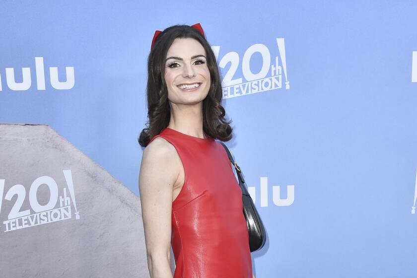 Dylan Mulvaney standing on red carpet smiling in red dress and red platform boots, carrying black hand bag