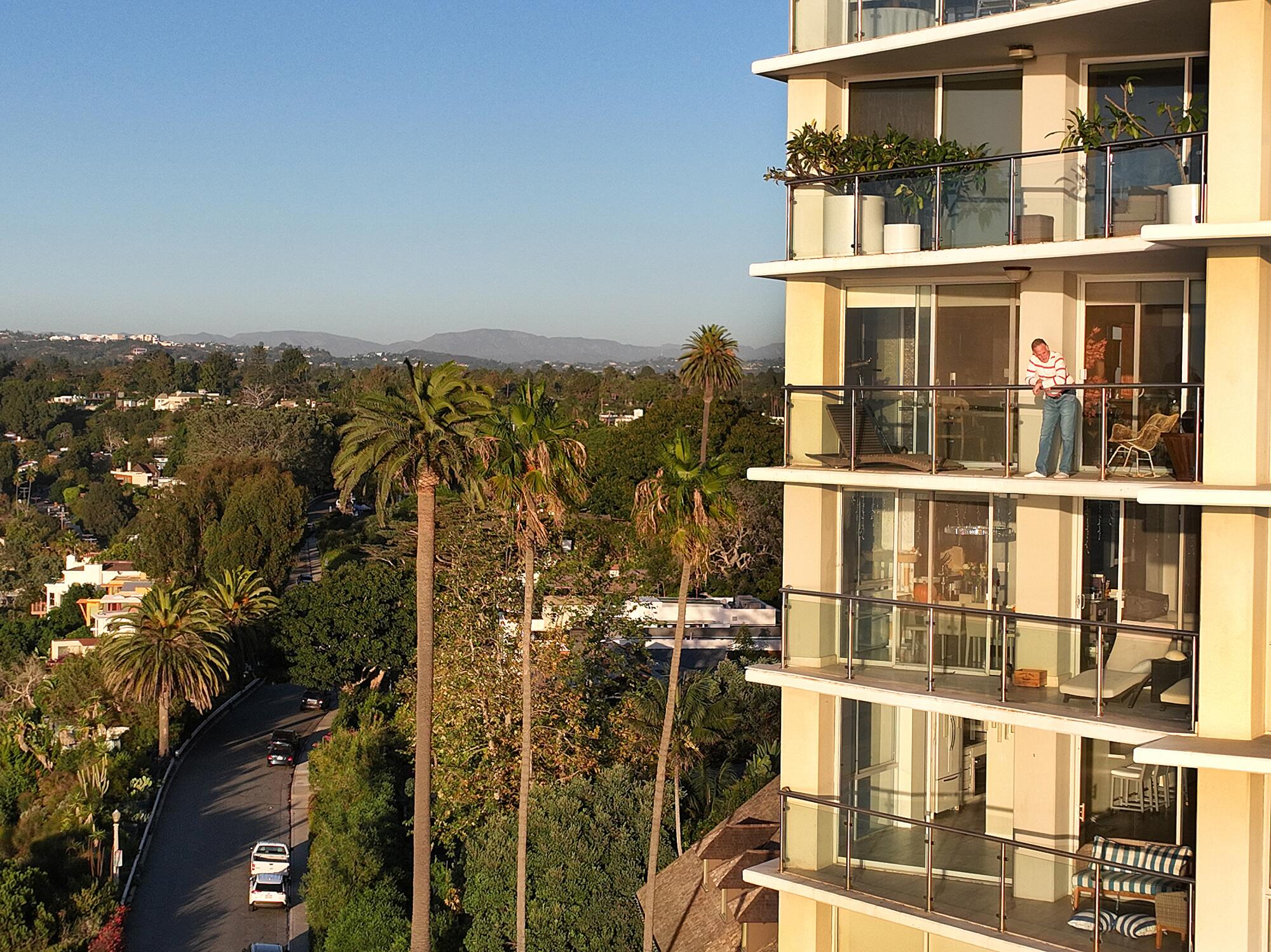 A person stands on a balcony of a residential tower above a tree-lined street.