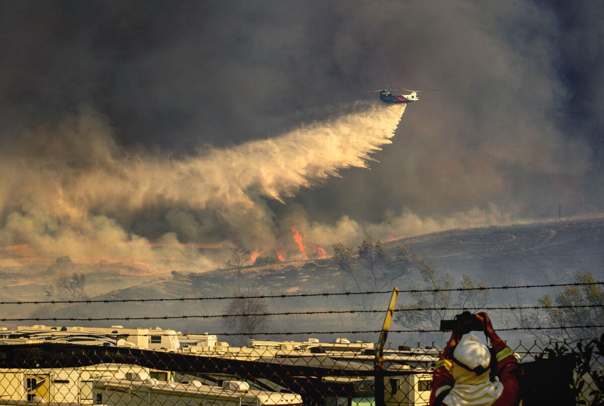 A person lifts a camera to record a helicopter as it drops water on leaping flames on a hillside.