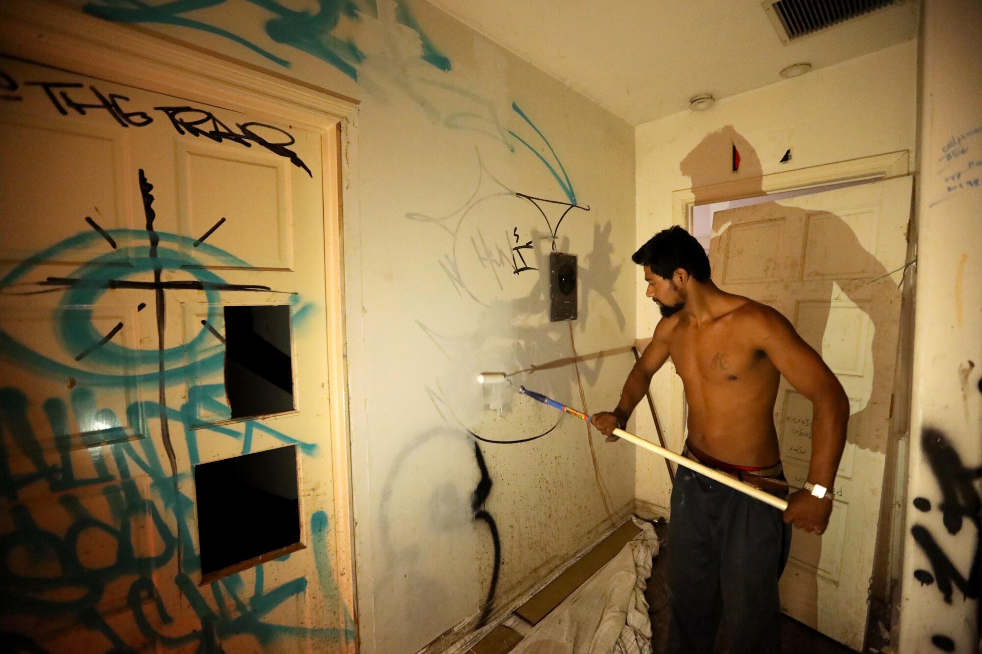 A shirtless man paints over graffiti inside a room