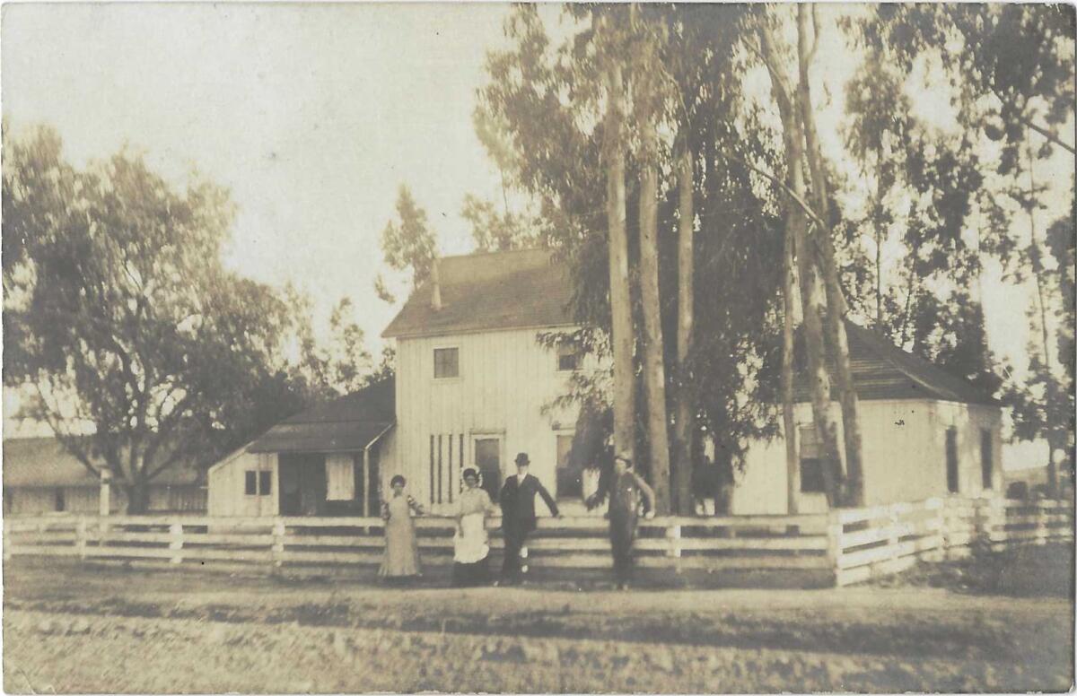 A sepia tone photograph shows two women and two men standing outside a farm house with a waist-high fence and mature trees