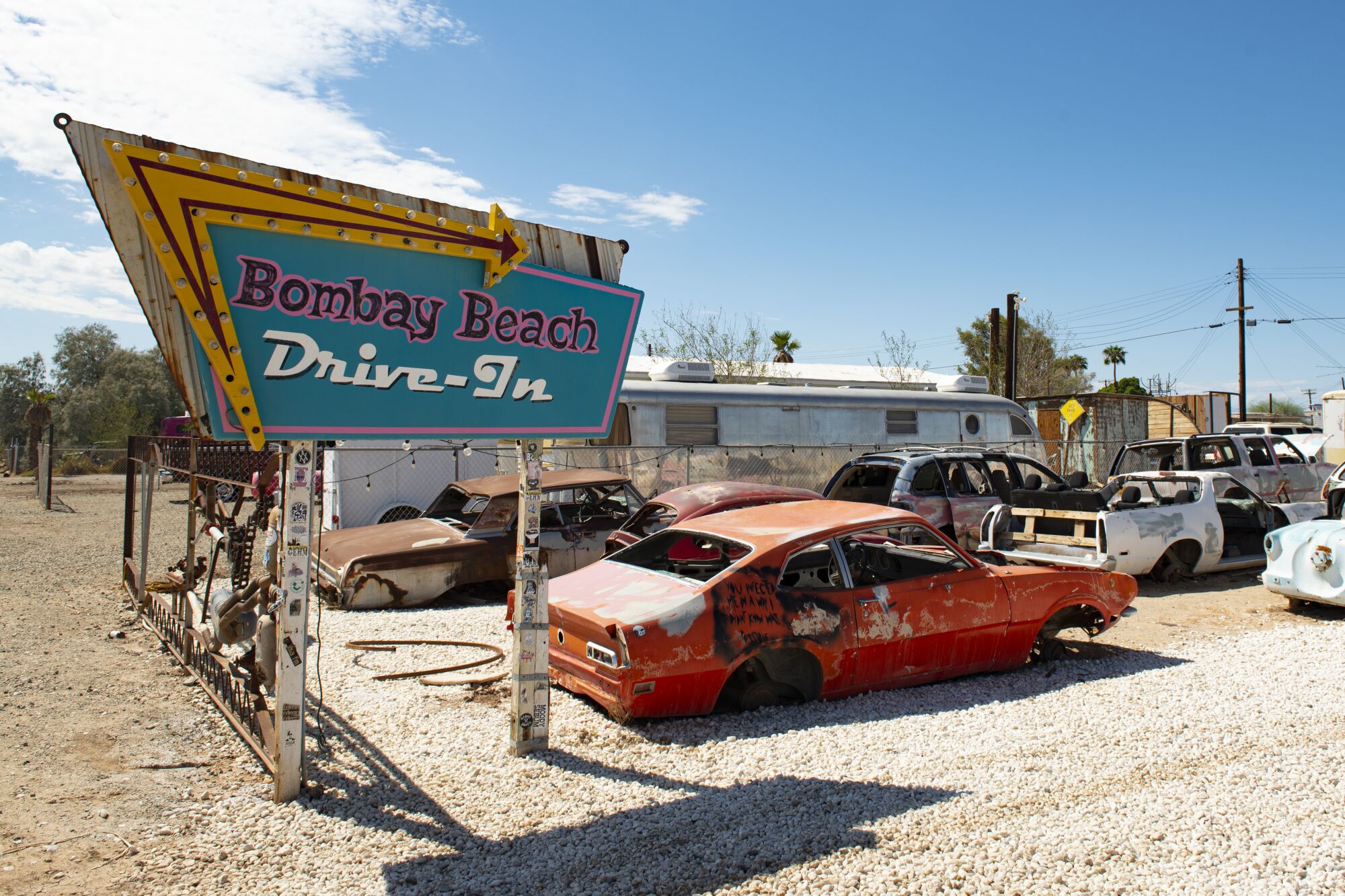 A sign that reads "Bombay Beach Drive-In" towers over dilapidated cars in a lot.