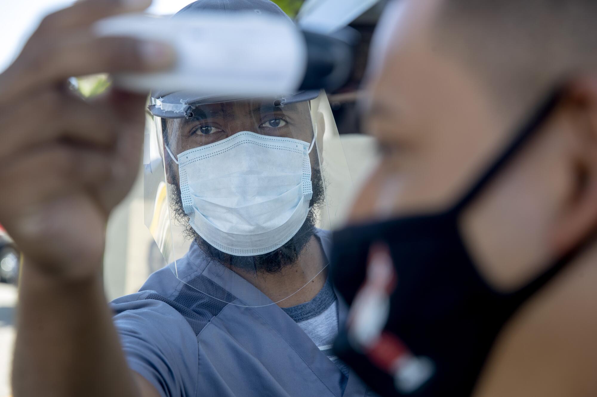 Nurse Bobby Binissa, wearing a mask and a plastic face shield, takes the temperature from a patient's forehead