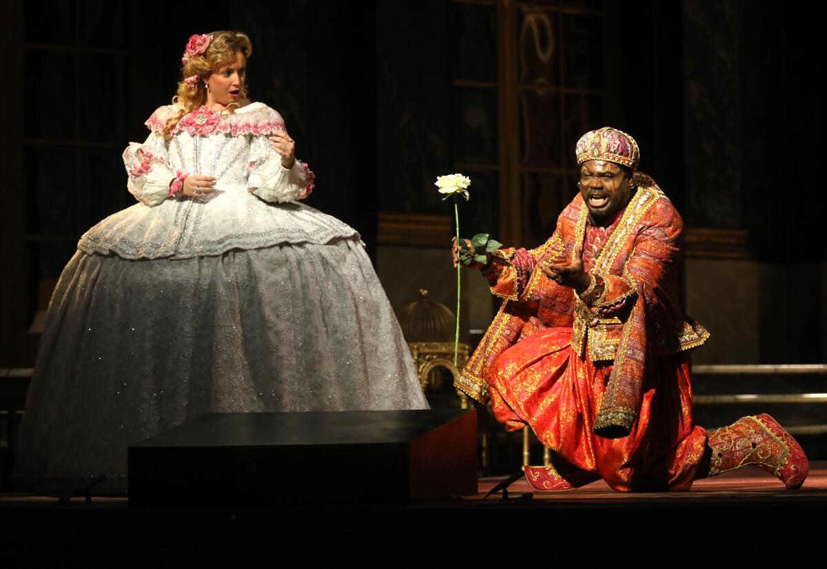 A Black man in elaborate costume sings on his knees next to a white woman in a 17th century gown in an opera scene.